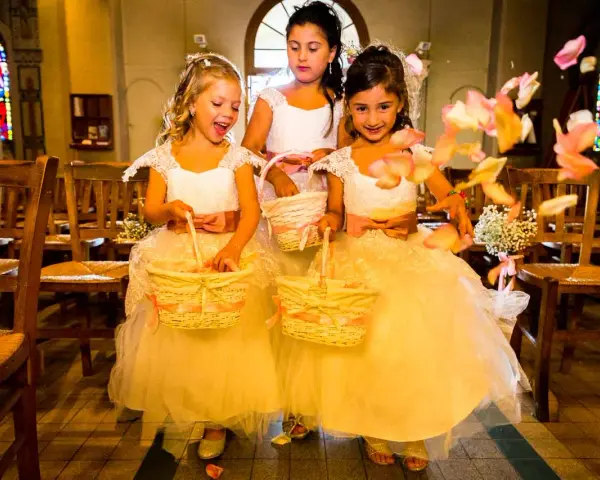 Trois enfants d'honneur souriantes, vêtues de robes claires, tiennent des paniers fleuris. Des pétales de roses volent autour d'elles dans une église.