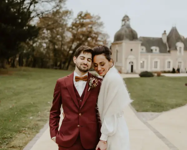 Un jeune marié rayonne dans son costume bordeaux, la couleur costume mariage parfaite pour un jour d'automne. Il enlace tendrement sa mariée devant un château.