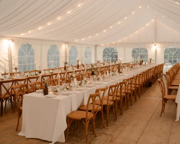 Ambiance chaleureuse sous tente pour un mariage, avec une longue table ronde ou rectangulaire dressée, éclairée par des guirlandes lumineuses et des bougies.