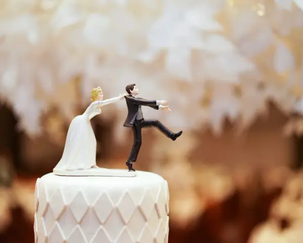 Figurines de mariés sur un gâteau. La mariée tire le marié par le revers. Un clin d'œil humoristique sur le prix d'un gâteau de mariage pour 100 personnes.