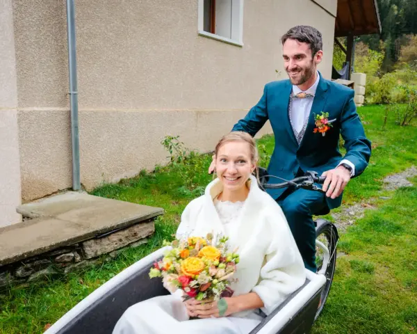 Un jeune couple souriant sur un vélo cargo, la mariée tenant un bouquet. Une idée originale pour un mariage avec un petit budget.