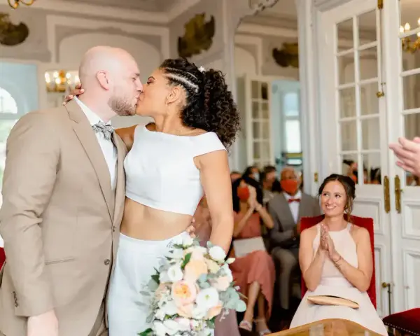 Un couple s'embrasse lors d'un mariage civil sans robe de mariée. La mariée porte un ensemble blanc moderne, le marié un costume beige.