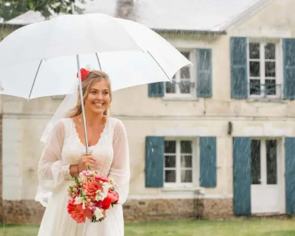 Mariée souriante sous un parapluie blanc, bouquet de fleurs à la main, devant une maison. Un mariage sous la pluie, mais le bonheur est là.