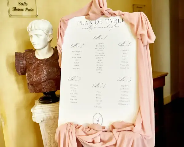 Plan de table de mariage, avec la disposition des invités par table. Une statue antique et des drapés roses décorent l'entrée.