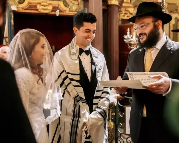 Un couple rayonne lors de leur mariage juif. Le marié, drapé d'un châle de prière, sourit à sa fiancée voilée. Un rabbin lit un document sacré.