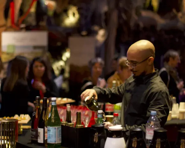 Un barman sert du vin lors d'un vin d'honneur pour 100 personnes. Des bouteilles et des verres sont disposés sur le comptoir.