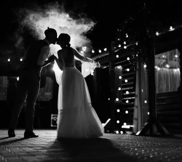 Silhouette d'un couple s'embrassant sous une fumée légère, éclairés par des guirlandes lumineuses. Une scène romantique qui évoque une chanson d'amour.