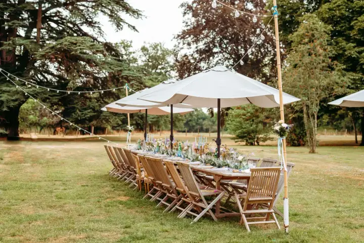 Table dressée pour un mariage jardin, avec des chaises en bois, des parasols et des guirlandes lumineuses.