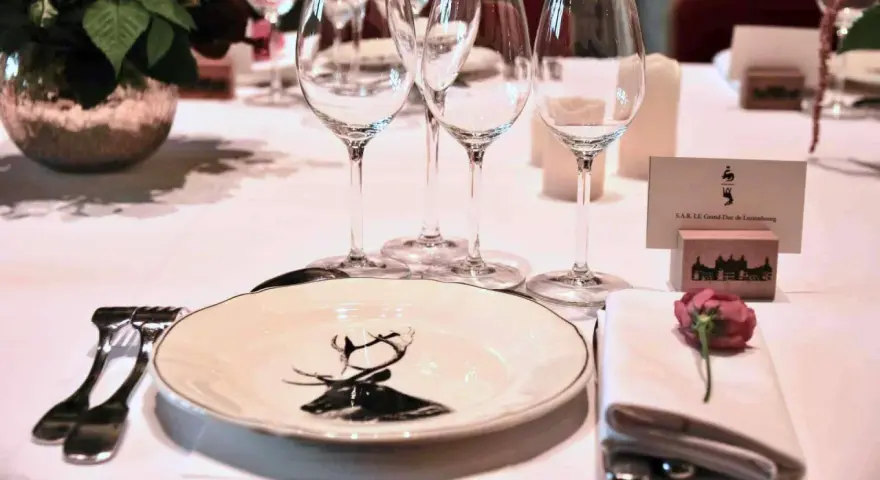 Une élégante table de mariage dressée avec des verres à pied, une assiette ornée d'un cerf, et un marque-place pour le Grand-Duc de Luxembourg.