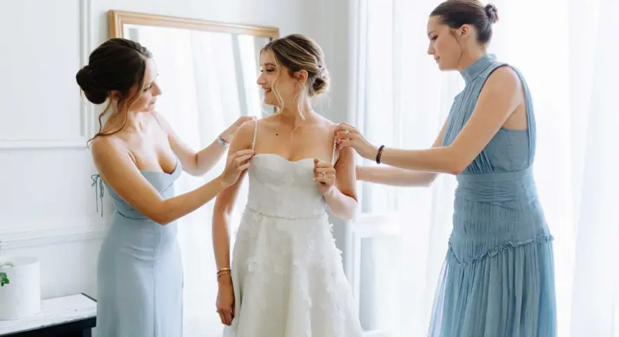 Trois femmes en robes bleues aident la mariée à enfiler sa robe blanche. Une question se pose : à quel âge peut-on être témoin de mariage ?