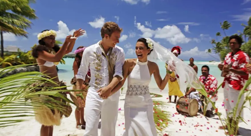Un jeune couple rayonnant célèbre son mariage à deux à l'étranger sur une plage tropicale, entouré de musiciens et de danseurs locaux.