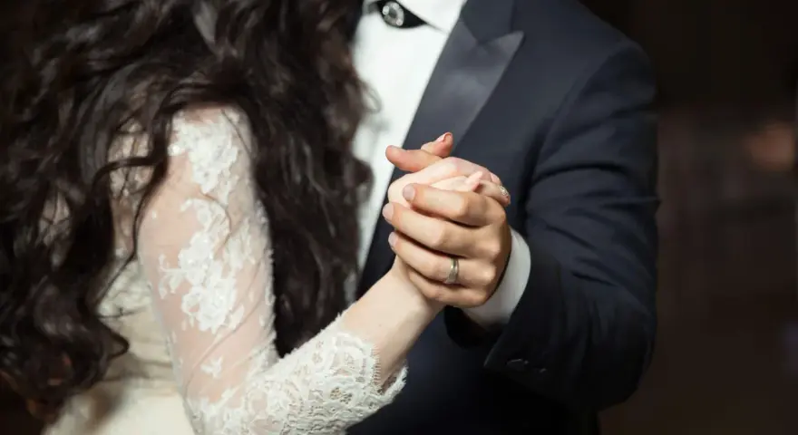 Les mains jointes d'un couple dansant une valse mariage. La mariée porte une robe en dentelle, le marié un costume sombre.