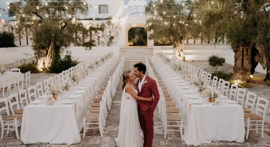 Un couple s'embrasse devant de longues tables dressées pour un mariage dans les Pouilles, illuminé par des guirlandes lumineuses.
