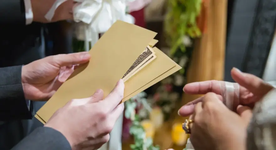 Une main tend une enveloppe dorée contenant des billets, un cadeau de mariage. Une autre main, ornée d'une bague, s'apprête à la recevoir.
