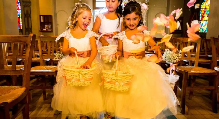Trois enfants d'honneur souriantes, vêtues de robes claires, tiennent des paniers fleuris. Des pétales de roses volent autour d'elles dans une église.