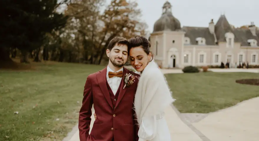 Un jeune marié rayonne dans son costume bordeaux, la couleur costume mariage parfaite pour un jour d'automne. Il enlace tendrement sa mariée devant un château.