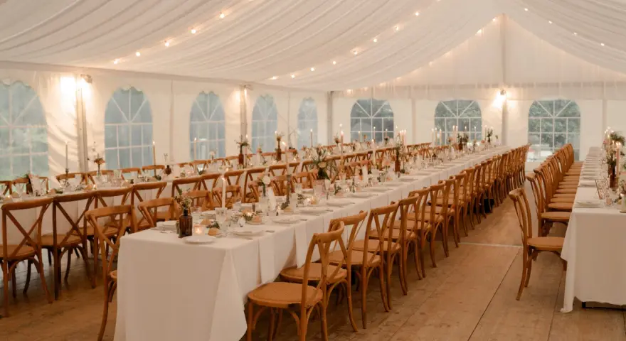 Ambiance chaleureuse sous tente pour un mariage, avec une longue table ronde ou rectangulaire dressée, éclairée par des guirlandes lumineuses et des bougies.