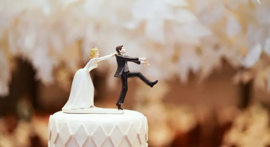 Figurines de mariés sur un gâteau. La mariée tire le marié par le revers. Un clin d'œil humoristique sur le prix d'un gâteau de mariage pour 100 personnes.