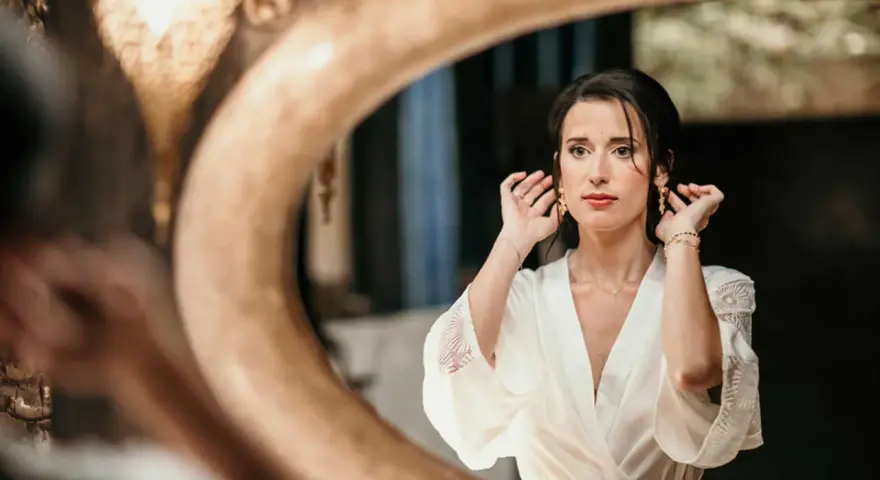 Une mariée, le visage marqué par le stress du mariage, ajuste ses boucles d'oreilles devant un miroir.