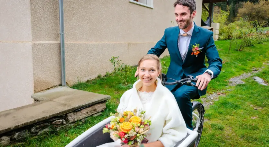 Un jeune couple souriant sur un vélo cargo, la mariée tenant un bouquet. Une idée originale pour un mariage avec un petit budget.