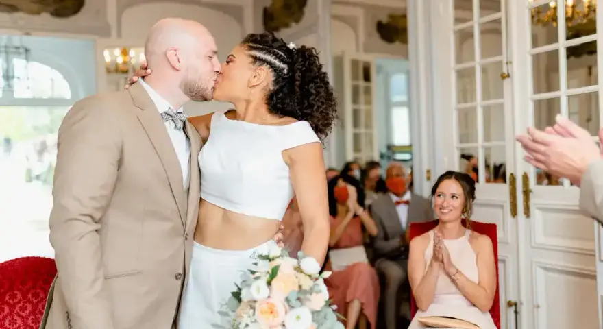 Un couple s'embrasse lors d'un mariage civil sans robe de mariée. La mariée porte un ensemble blanc moderne, le marié un costume beige.