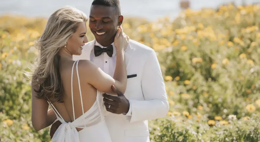 Un couple heureux dans un champ de fleurs jaunes. La mariée porte une robe de mariée blanche, simple et élégante, avec un dos nu. Le marié est en costume blanc.