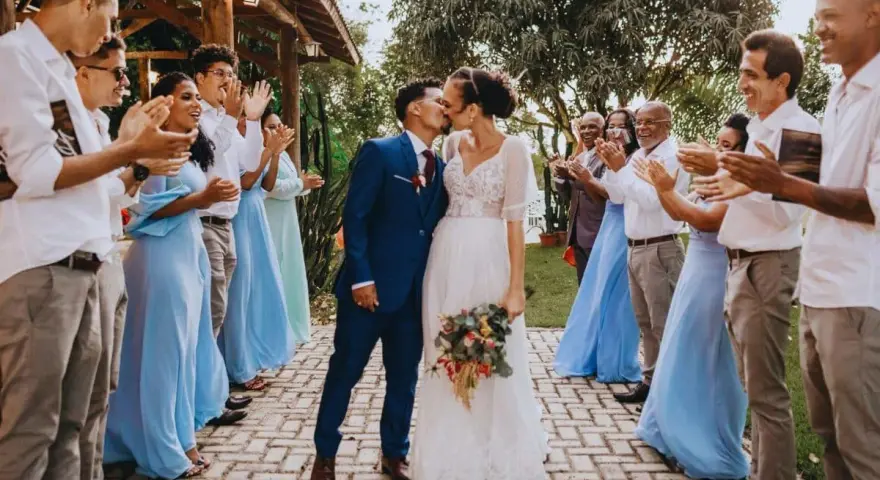 Un couple s'embrasse lors de leur petit mariage de 20 personnes. Les invités, en tenues bleues et blanches, applaudissent joyeusement.