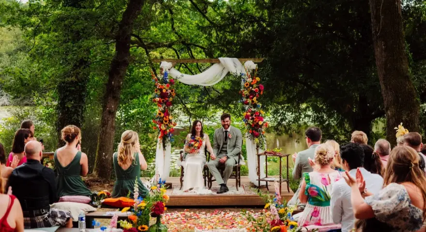 Un couple souriant lors d'une cérémonie laïque en plein air, entouré de verdure et de fleurs colorées. L'officiant cérémonie laïque guide ce moment unique.