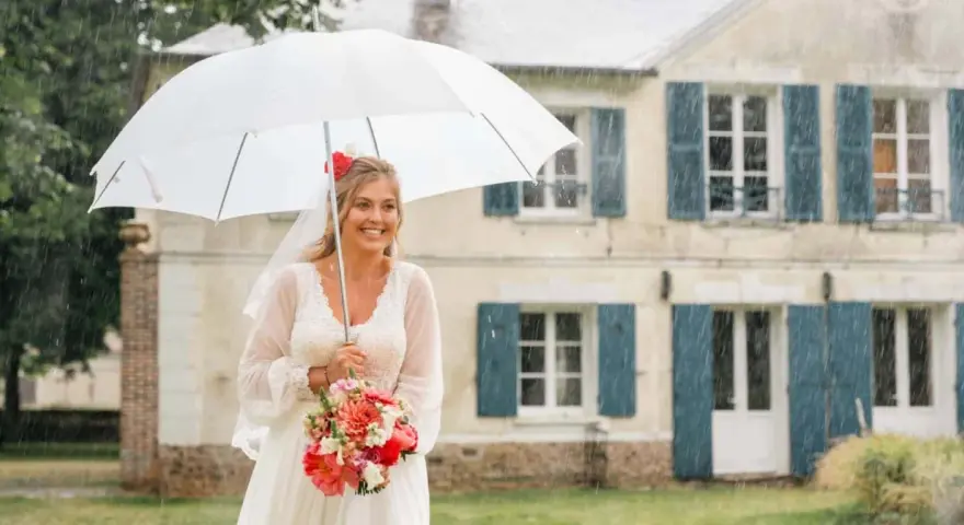 Mariée souriante sous un parapluie blanc, bouquet de fleurs à la main, devant une maison. Un mariage sous la pluie, mais le bonheur est là.