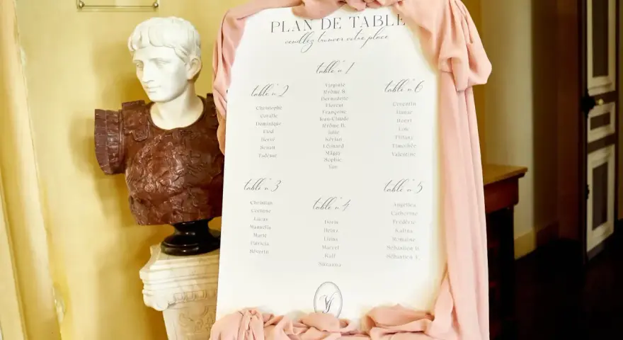 Plan de table de mariage, avec la disposition des invités par table. Une statue antique et des drapés roses décorent l'entrée.