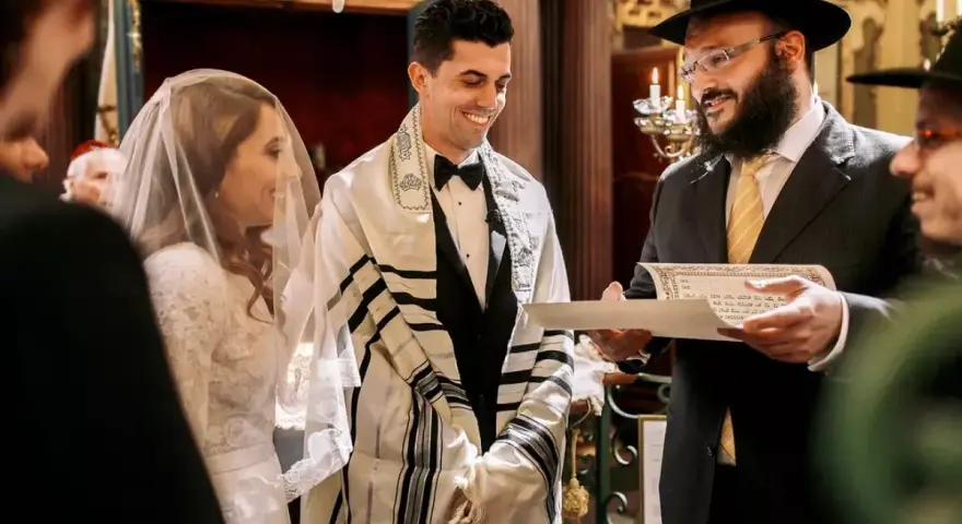 Un couple rayonne lors de leur mariage juif. Le marié, drapé d'un châle de prière, sourit à sa fiancée voilée. Un rabbin lit un document sacré.