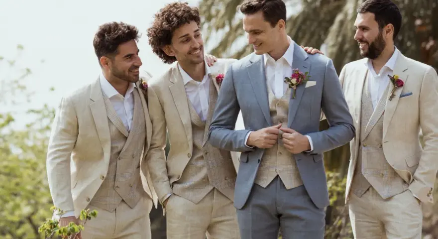 Quatre hommes en costumes de lin, dont un témoin et un garçon d'honneur, sourient et discutent à l'extérieur.