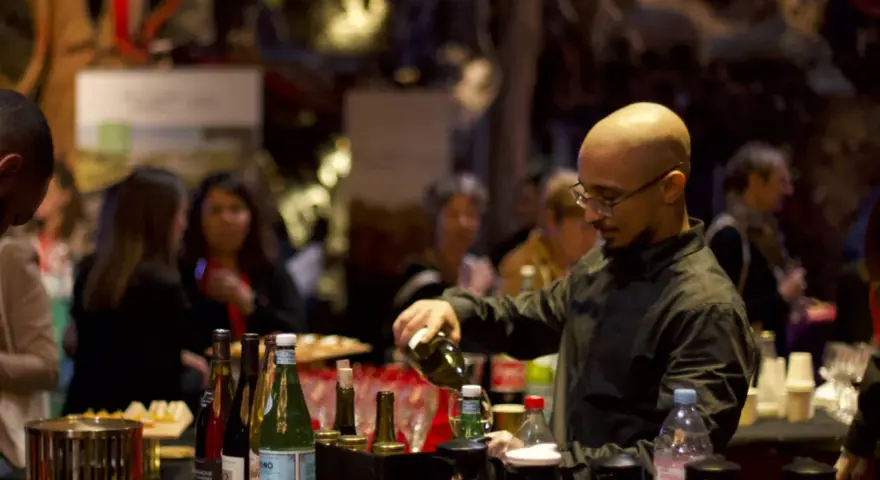 Un barman sert du vin lors d'un vin d'honneur pour 100 personnes. Des bouteilles et des verres sont disposés sur le comptoir.