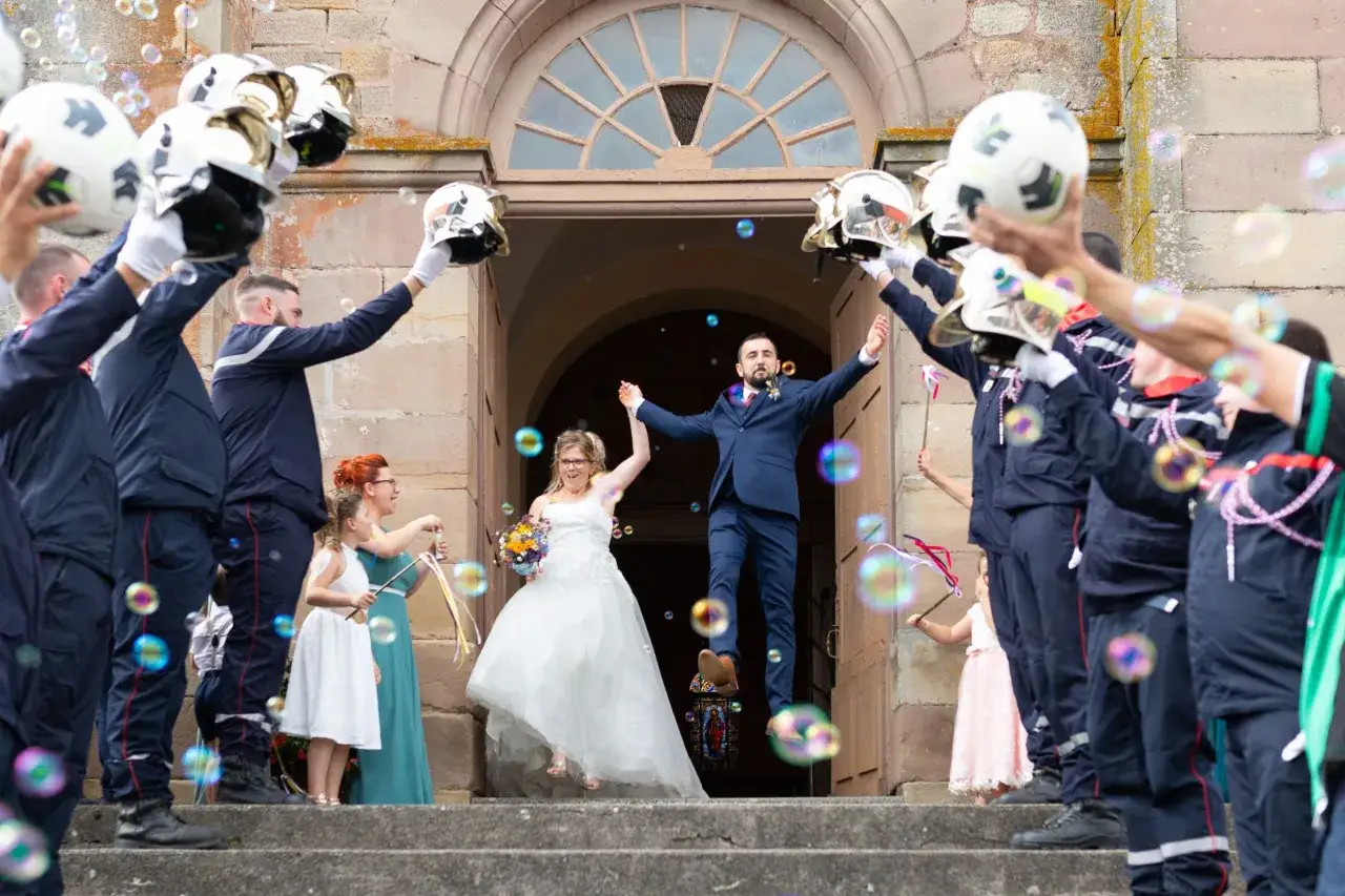 Mariage joyeux : les mari&eacute;s sortent de l'&eacute;glise sous une haie d'honneur de pompiers lan&ccedil;ant des bulles.