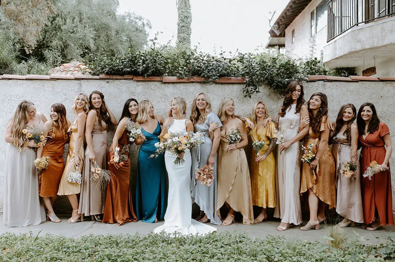 Un groupe de demoiselles d'honneur souriantes, vêtues de robes aux couleurs vives, pose devant un mur en pierre.