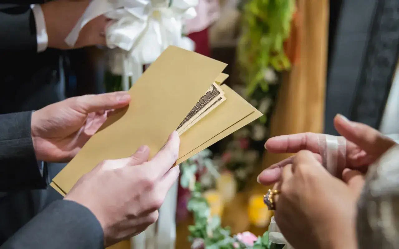 Une main tend une enveloppe dorée contenant des billets, un cadeau de mariage, à une autre main ornée d'une bague.