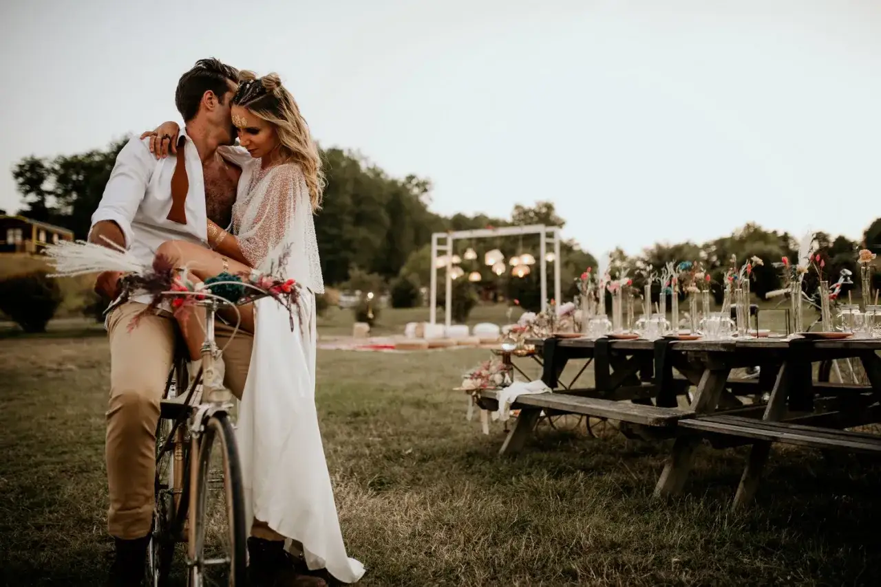 Un couple s'embrasse sur un vélo décoré, devant une table dressée pour un mariage champêtre.