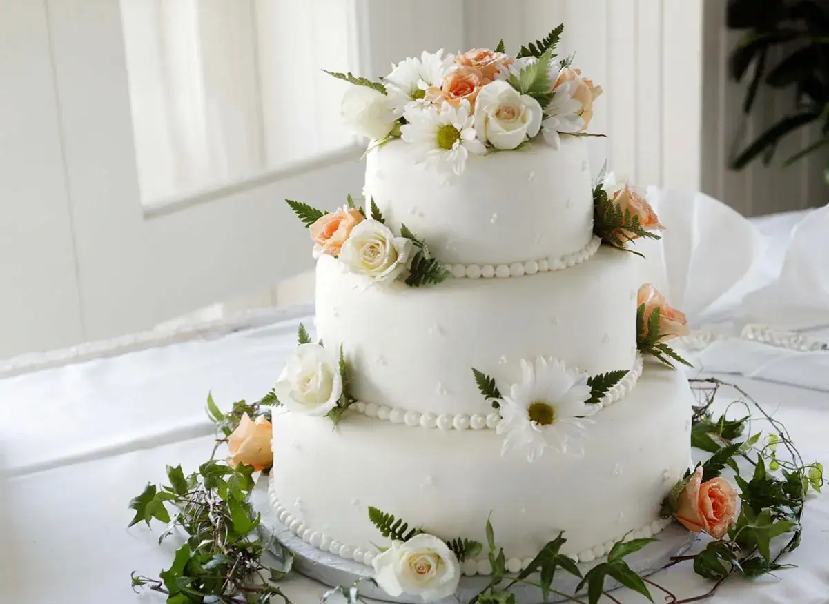 Magnifique gâteau de mariage traditionnel à trois étages, orné de roses et de marguerites, avec des guirlandes de verdure.