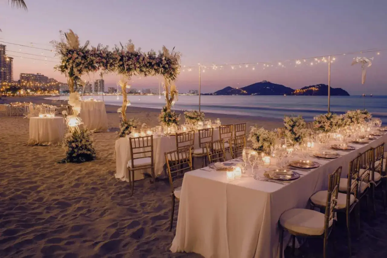 Un décor de mariage insolite sur la plage au crépuscule. Tables dressées, fleurs, bougies et guirlandes lumineuses créent une ambiance féerique.