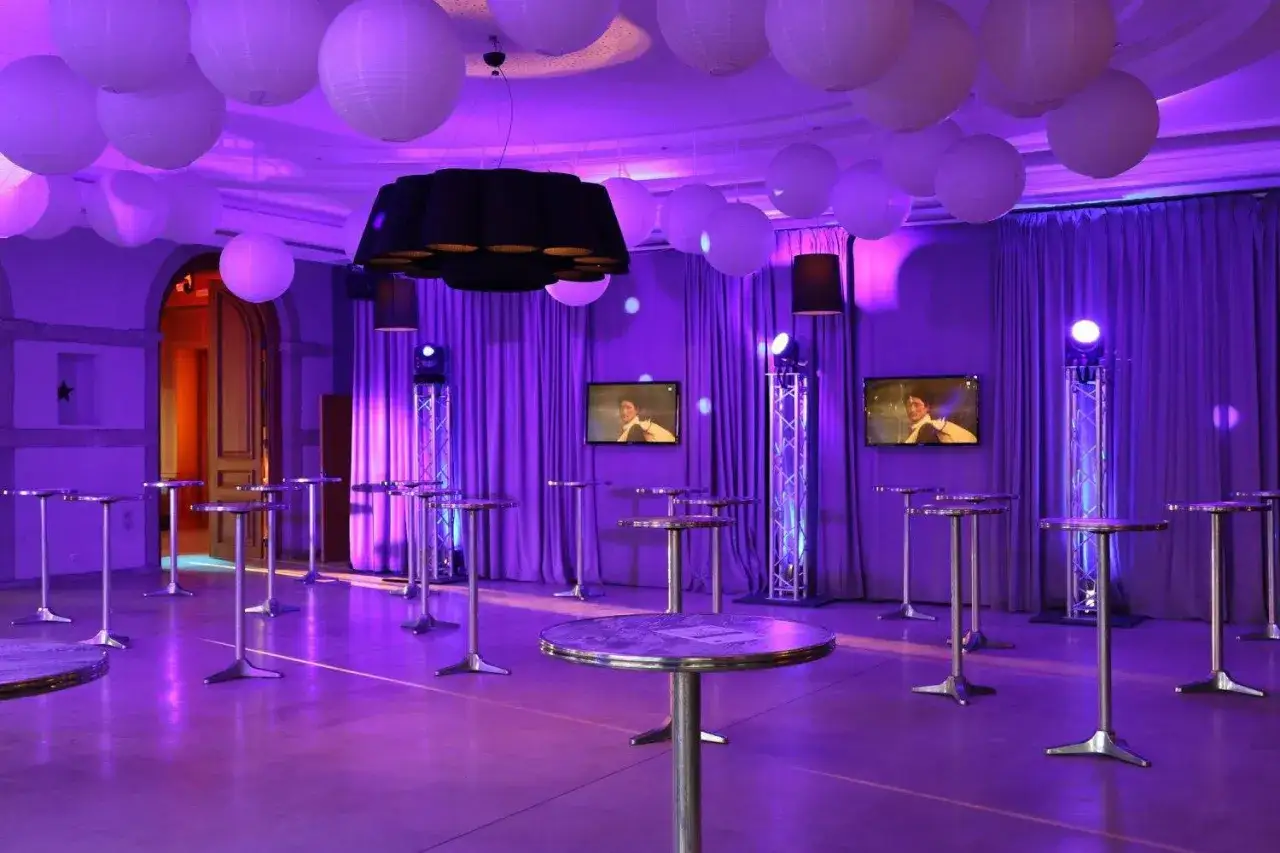 Salle de réception violette avec des lanternes blanches, des tables hautes et un totem jeux de lumière diffusant une ambiance festive.