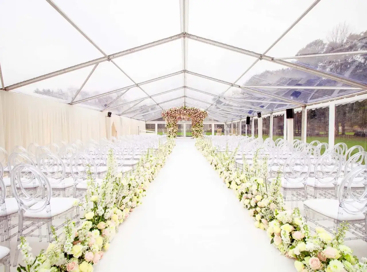 Allée de mariage sous tente transparente, décorée de fleurs roses et blanches. Parfait pour un mariage pluie.