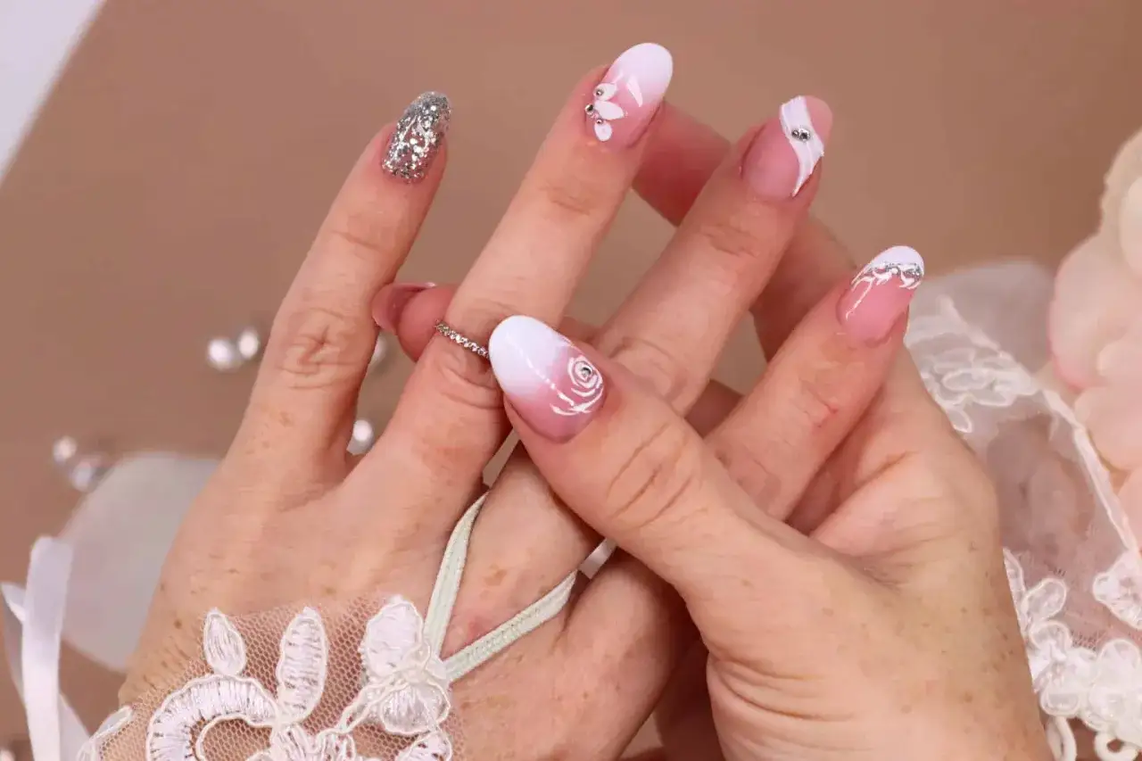 Manucure délicate pour un mariage bohème : ongles dégradé blanc et rose, ornés de roses, de strass et de paillettes argentées.