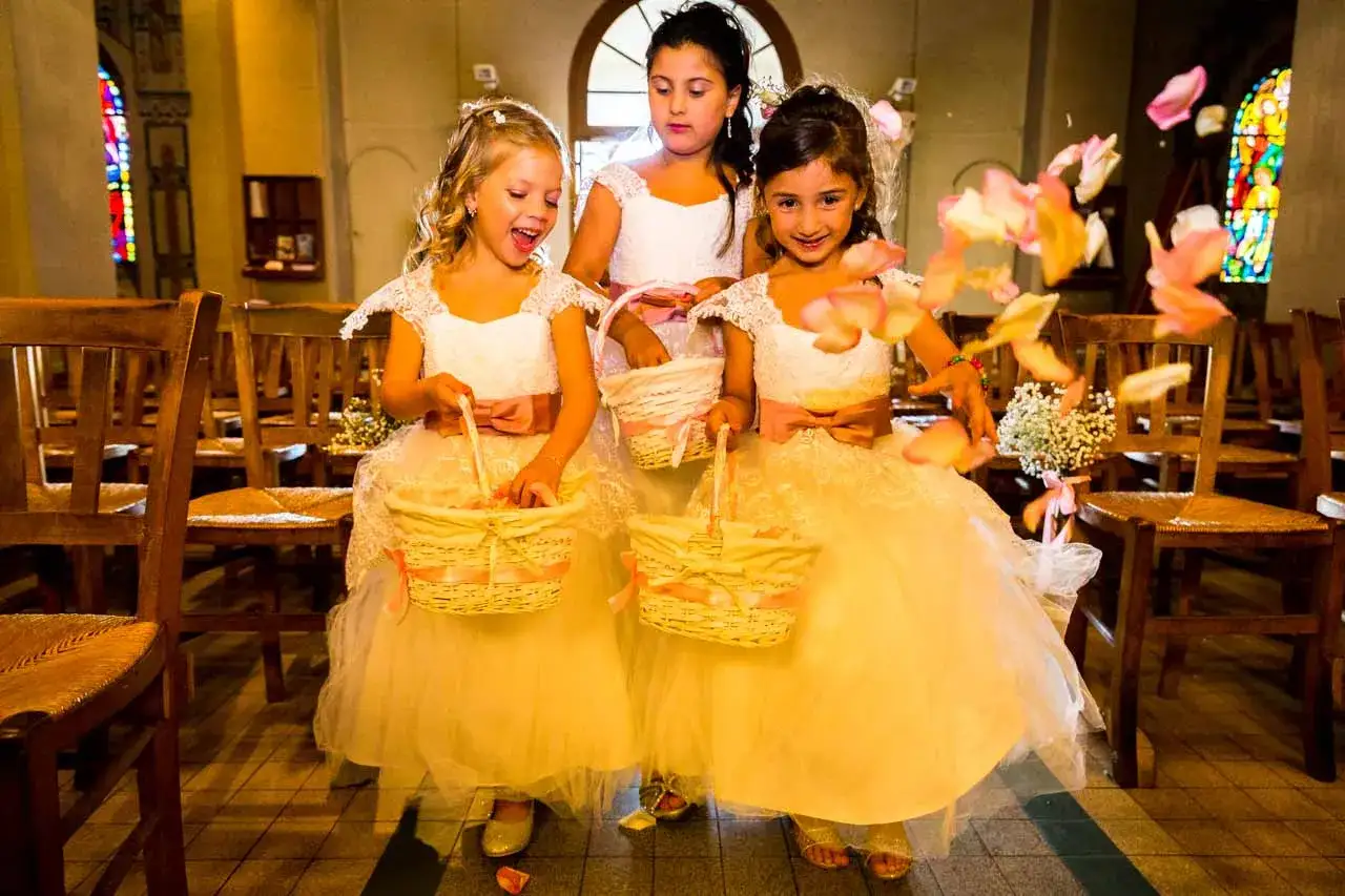 Trois enfants d'honneur souriantes, vêtues de robes blanches et dorées, tiennent des paniers en osier remplis de pétales de roses.