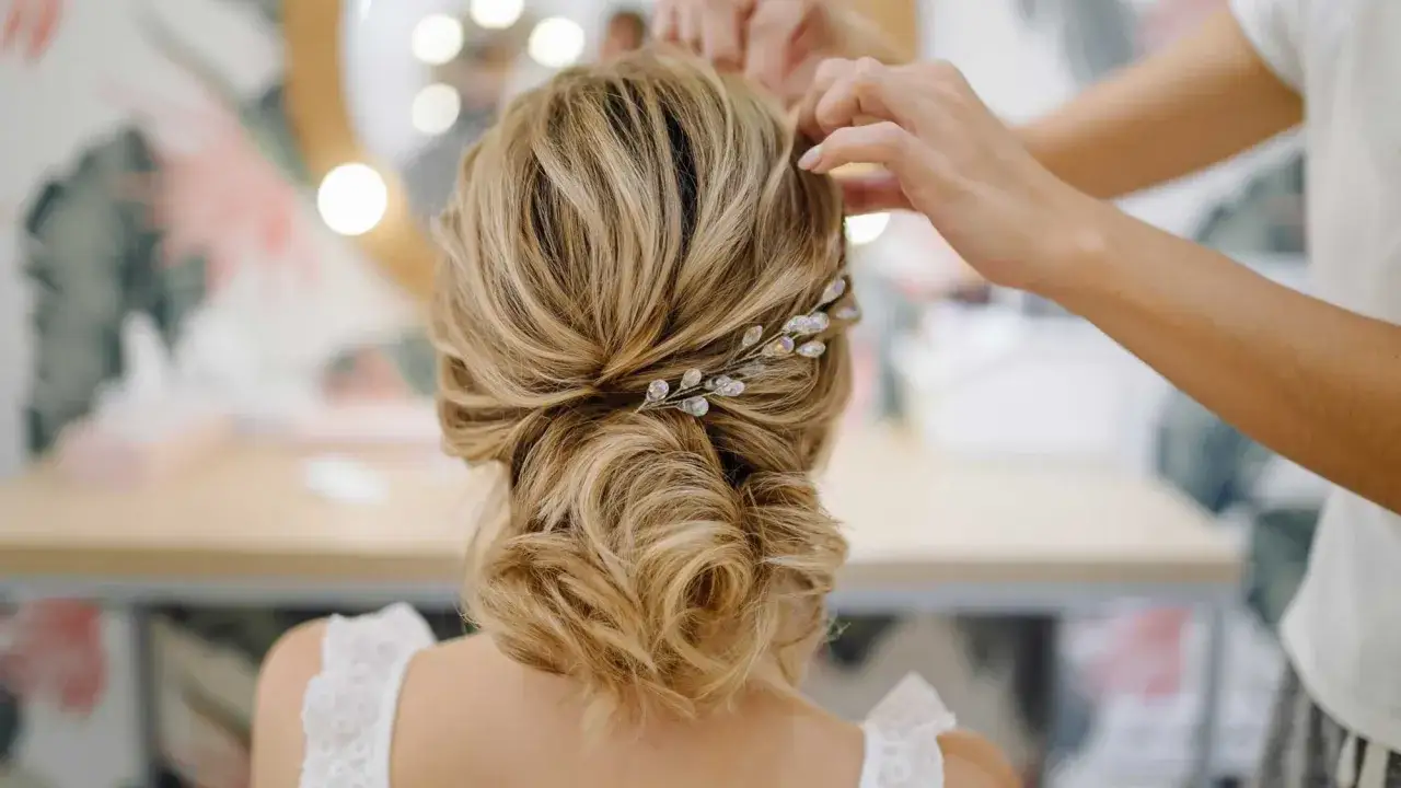 Chignon mariage romantique aux boucles souples, orné d'une délicate barrette scintillante. Les mains d'une coiffeuse finalisent la mise en beauté.