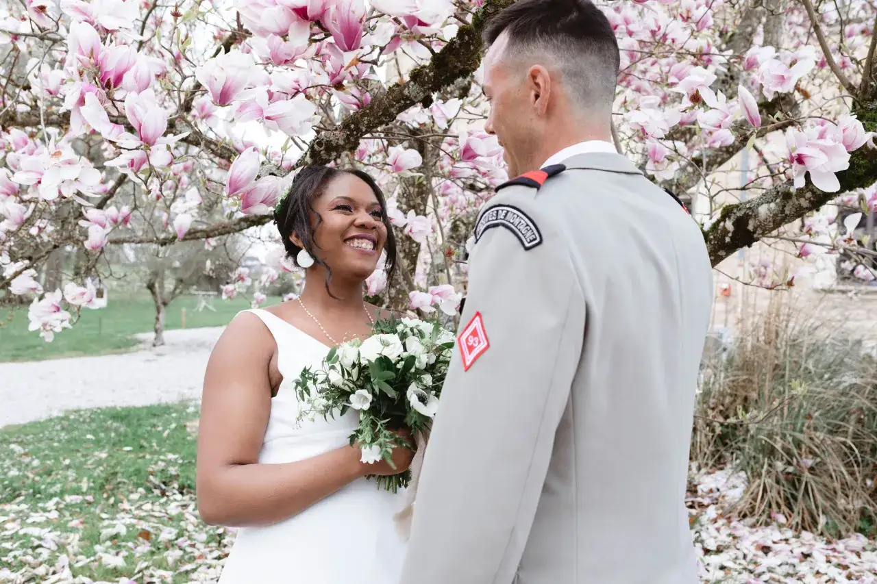 Un couple rayonnant lors de leur mariage militaire, sous un cerisier en fleurs. La mariée, radieuse, tient un bouquet blanc.