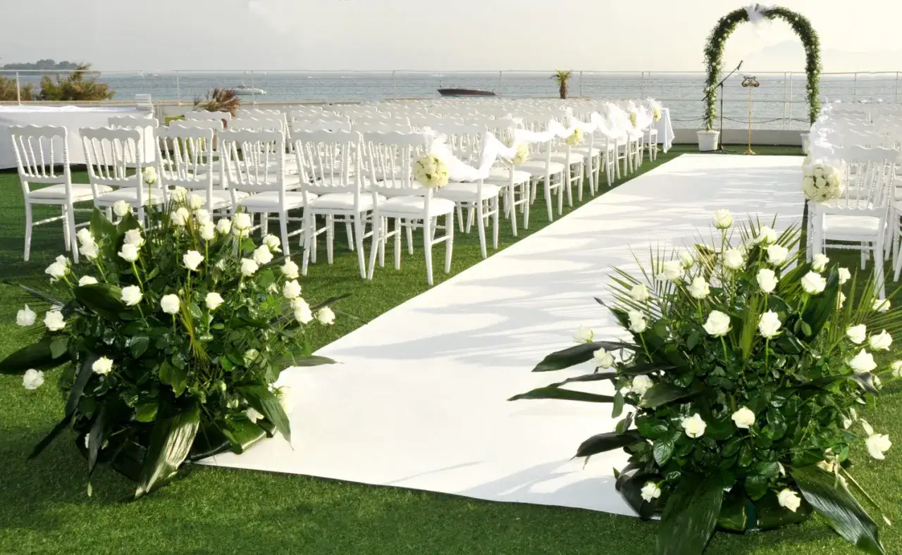 Aménagement d'une cérémonie laïque avec chaises blanches, tapis, arche fleurie et bouquets de roses. L'organisation est soignée pour un événement mémorable en bord de mer.