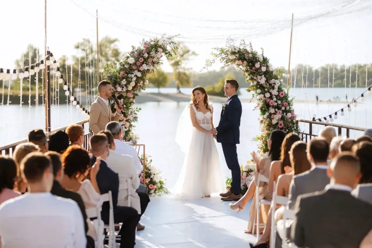 Un officiant de cérémonie laïque guide un mariage en plein air, sous une arche fleurie, avec les mariés et leurs invités.