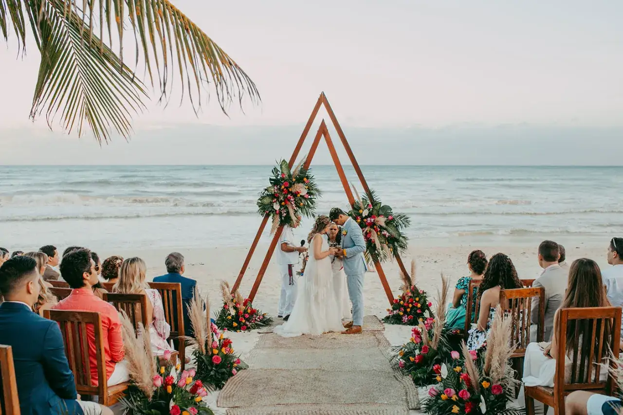 Cérémonie de mariage à deux à l'étranger sur une plage tropicale, avec un arche décoré de fleurs, des invités assis et l'océan en arrière-plan.