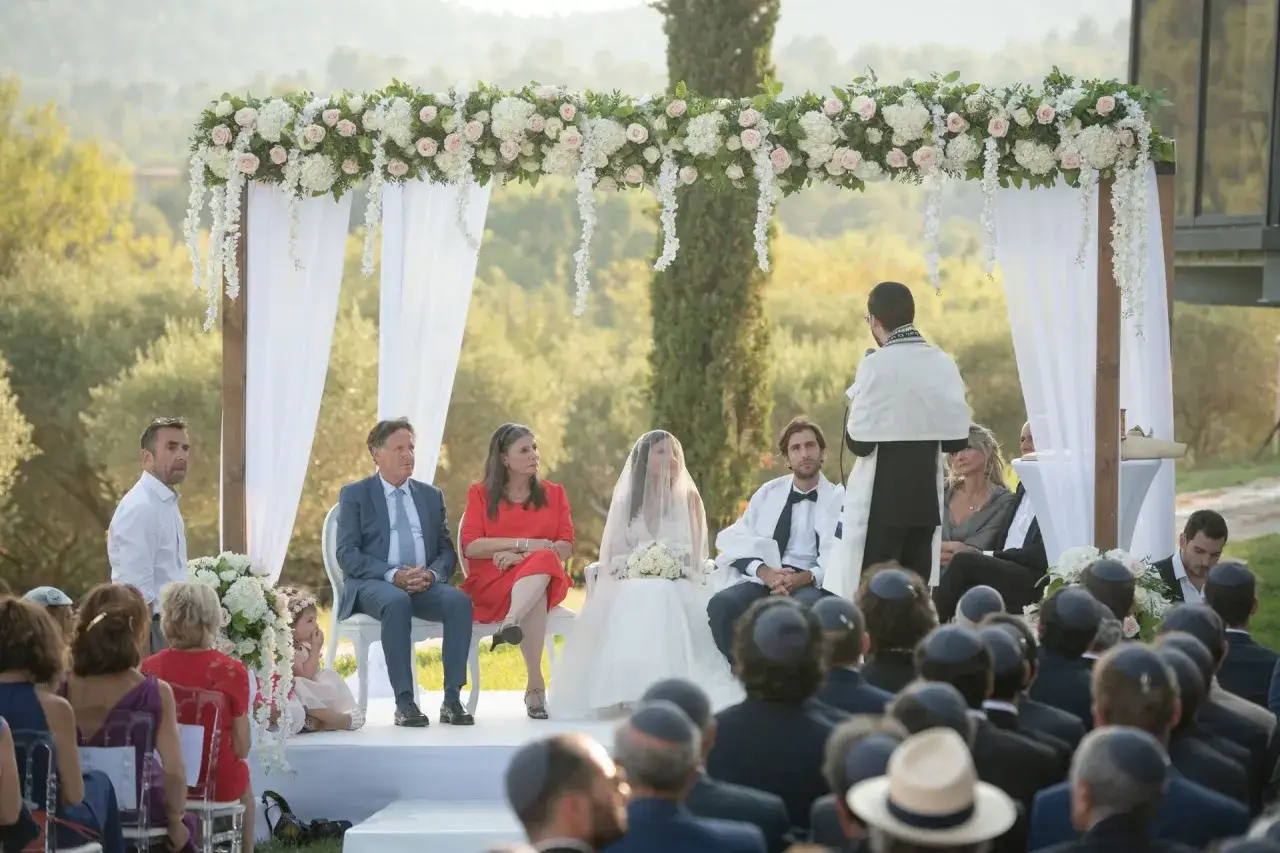 Cérémonie de mariage juif sous une houppa fleurie. La mariée, voilée, est assise avec ses proches. L'officiant s'adresse aux invités.