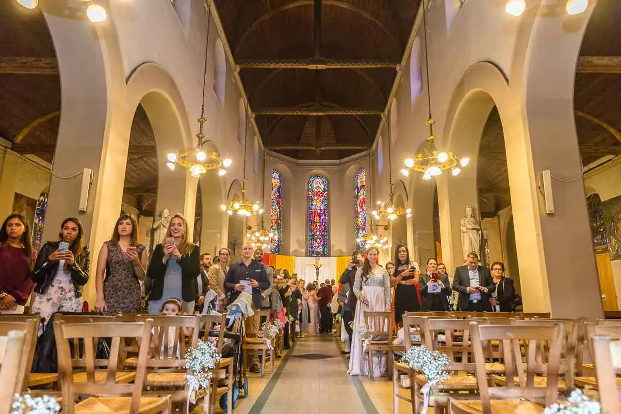 Des invités attendent dans une église, certains filmant le cortege mariage avec leurs téléphones.